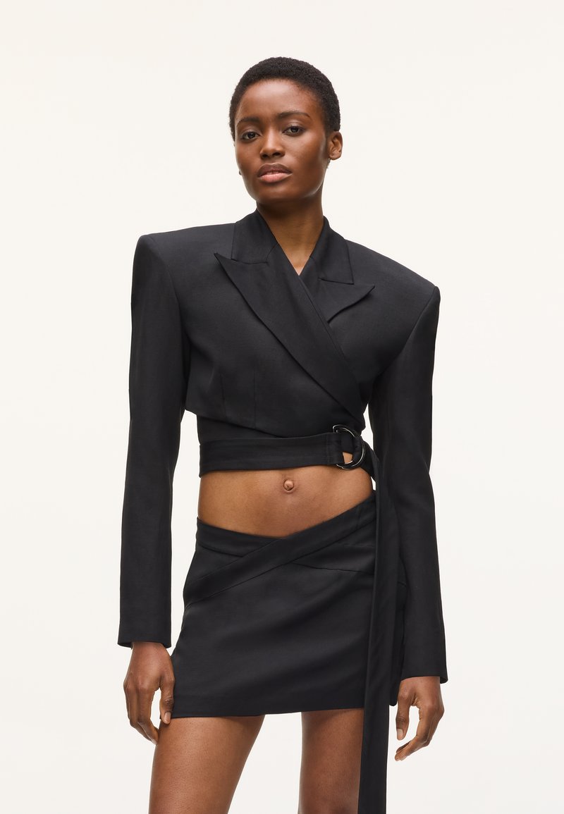Γυναίκα που φοράει ένα μαύρο cropped σακάκι με φαρδιούς ώμους και ζώνη στη μέση, συνδυασμένο με μια μίνι ασύμμετρη φούστα που ταιριάζει.