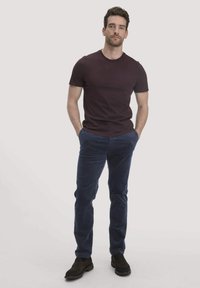 Bordeaux katoenen t-shirt gecombineerd met marineblauwe corduroy broek. Het model staat met de handen in de zakken en draagt donkere schoenen. Eenvoudige, casual pasvorm.
