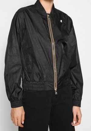 Veste bomber légère noire avec poignets et ourlet élastiques, fermeture éclair avant avec bordure orange et blanche, portée par une personne en pantalon noir.