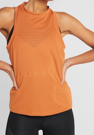 Gros plan d’une personne portant un haut de sport sans manches orange perforé et un legging noir, les mains posées sur les hanches.