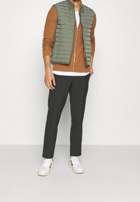 Green quilted vest over a rust-colored sweater, paired with dark, tapered trousers and white sneakers with black accents.