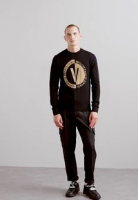 Zwarte lange mouwen trui met een groot gouden "V" logo op de voorkant, gecombineerd met zwarte cargo-broeken en zwarte sneakers. Glad materiaal.