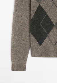 Manica di un maglione marrone in lana con motivo a rombi argenteo scuro e nero e polsino a costine su sfondo bianco.