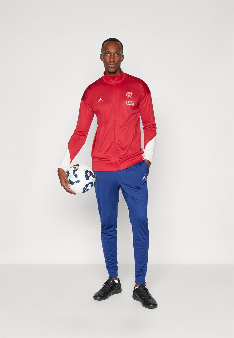 Chaqueta de entrenamiento roja con detalles y logotipo en blanco, combinada con pantalones azules. La figura sostiene un balón de fútbol con estampado y lleva zapatillas negras.