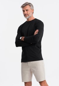 Schwarzes Langarmshirt mit rundem Ausschnitt, kombiniert mit hellbeigen Shorts; Modell trägt eine Armbanduhr und steht mit verschränkten Armen.