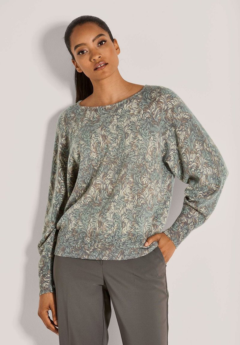Vrouw met donker haar in een paardenstaart draagt een lange mouw, een gemêleerde groene en beige trui en een grijze broek, poseert met één hand in haar zak.