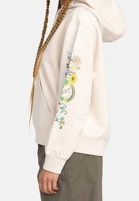 Personne portant un sweat à capuche beige avec des motifs floraux colorés et un arc-en-ciel sur la manche ainsi qu'un pantalon vert olive, debout devant un fond uni.
