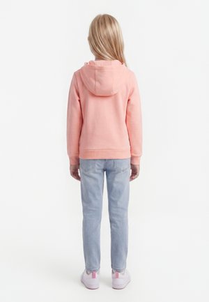 Dieťa s dlhými blond vlasmi stojace chrbtom, oblečené v ružovej mikine s kapucňou, svetlomodrých džínsoch a bielych teniskách s ružovými akcentmi.