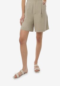 Pantalones cortos beige de tejido ligero, con cintura alta, pliegues y un corte relajado. Combinados con sandalias deslizantes en un tono pálido.