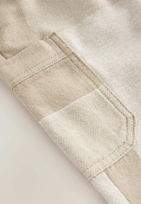 Beigefarbener Stoff mit strukturiertem Finish, ausgestattet mit einer Seitentasche und kontrastierenden Stoffteilen. Glattes, robustes Material ist sichtbar.