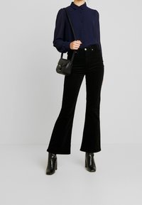 Blouse bleu marine à manches longues avec des boutons, associée à un pantalon en velours côtelé noir évasé et des bottines à talons noires, tenant un petit sac noir.