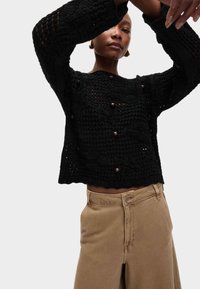 Pull en tricot noir avec un motif texturé et des boutons marron discrets, associé à un pantalon beige taille haute. La coupe est décontractée.