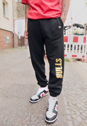 Pantalones de chándal negros con "BULLS" en amarillo en la pierna izquierda, combinados con zapatillas blancas y negras con acentos rojos.