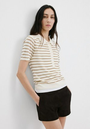 Žena s dugom tamnom kosom u kremasto bijeloj polo majici kratkih rukava s tankim smeđim prugama i crnim šortsom, stoji uz bijeli zid.