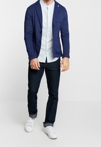 Námořnický blejzr přes světle modrou košili na knoflíky, spárovaný s tmavými džíny a bílými teniskami. Blejzr má dva knoflíky a náprsní kapsu.