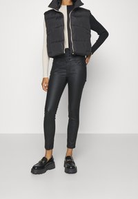 Person med svart pufferväst, beige och svart långärmad topp, svarta högmidjade byxor och svarta klumpiga loafers står mot en vit bakgrund.