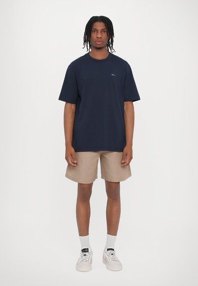 T-shirt en coton marine à manches courtes, associé à un short beige. Chaussures de sport de couleur claire avec lacets et chaussettes blanches mi-mollet.