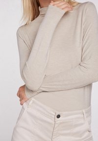 Beige pidähuppari, jossa on pitkät hihat, valmistettu pehmeästä ja kevyestä kankaasta. Yhdistetty kerma värisiin housuihin, joissa on nappiyksityiskohta.