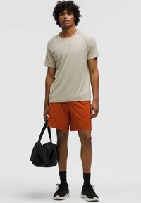 Ljusbeige texturerad t-shirt i kombination med orange shorts, svarta sportstrumpor och sneakers, håller en svart duffelväska.