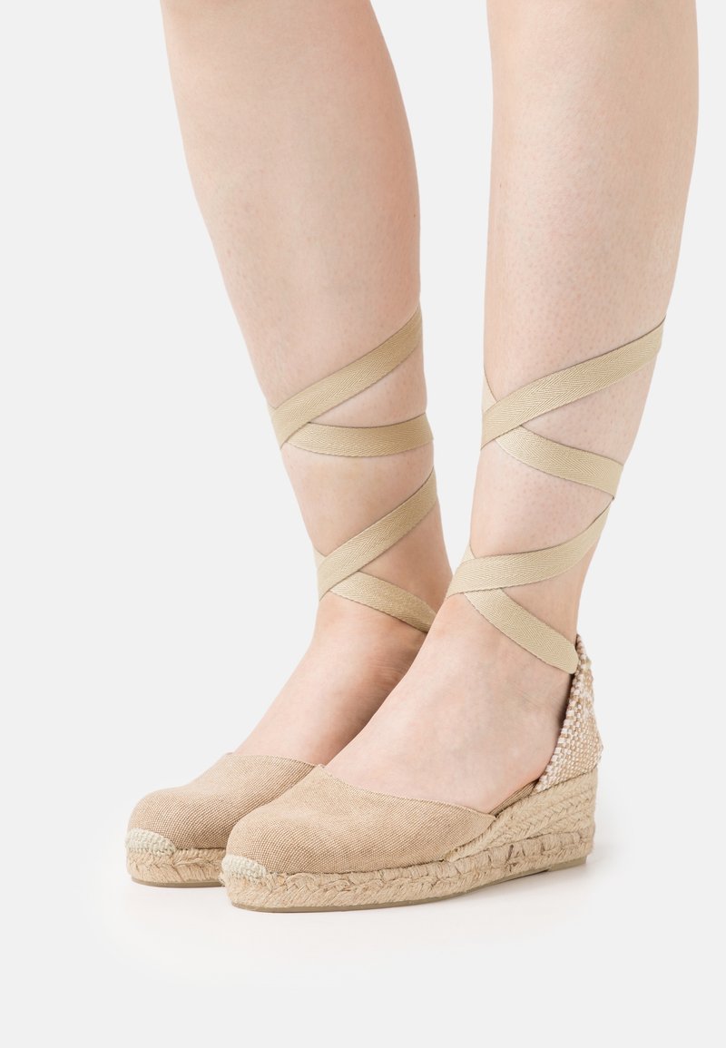 Chaussures compensées espadrilles beige avec rubans à nouer autour de la cheville sur jambes pâles, fond blanc