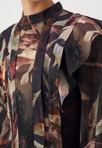 Blouse florale transparente avec col haut et détail froncé à l'épaule dans des couleurs sombres et atténuées avec des panneaux de tissu verticaux.