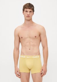 Jonge man met kort bruin haar, staande, gekleed in gele Nike boxershorts, op een effen witte achtergrond.