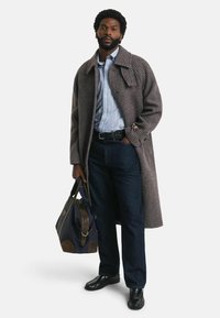 Abrigo de espiga gris, camisa de rayas azul claro, jeans oscuros, zapatos negros, sosteniendo una bolsa azul oscura con detalles en marrón.