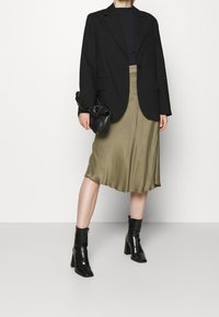 Blazer noir cintré porté sur un haut bleu marine, associé à une jupe en satin olive lisse. Bottines noires à talon carré et petit sac noir.