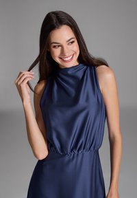 Robe en satin bleu marine avec un col haut, sans manches et une taille froncée. Texture lisse avec une silhouette fluide.
