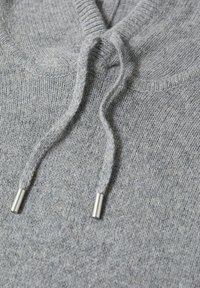 Szara bluza z kapturem o fakturowanym wzorze dzianiny, z troczkiem z metalowymi srebrnymi końcówkami oraz ściągaczem przy dekolcie. Miękki, casualowy materiał.