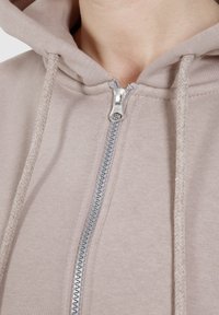 Lichtbeige ritsvest met een zilveren rits, koordjes en een textuurstof. Close-up van de kraag en het ritsdetail.