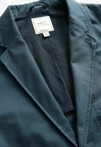 Blazer su misura blu scuro con colletto a punta, realizzato in tessuto liscio. Presenta un'etichetta "NEXT" con "Regular Fit" e istruzioni per la cura all'interno.