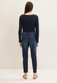 Mujer de pie, descalza, vistiendo una camiseta de manga larga oscura y unos jeans oscuros ajustados, mirando hacia una pared lisa de color claro.