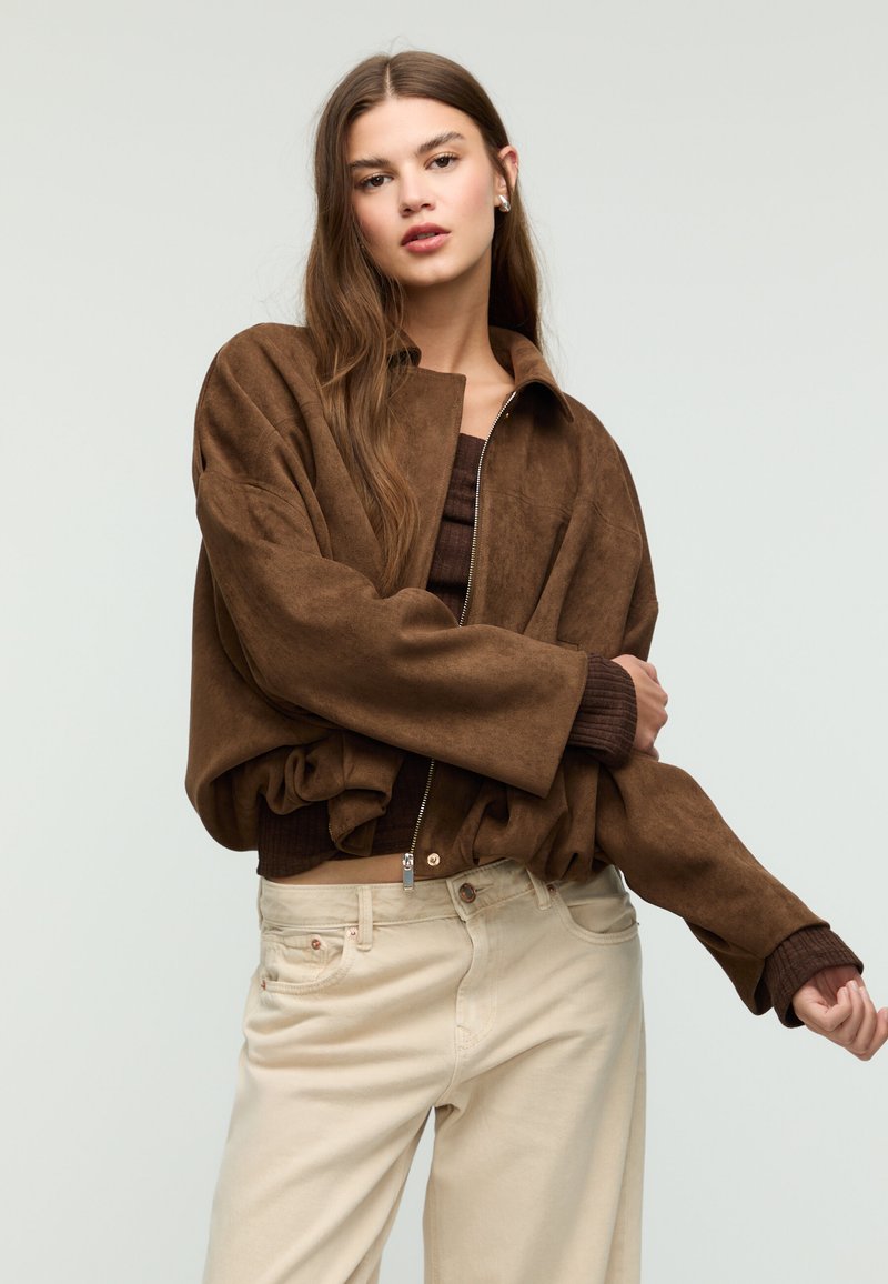 Giacca marrone in finta pelle scamosciata con zip frontale e maniche larghe, indossata sopra una maglia marrone a coste e pantaloni beige. Texture liscia, vestibilità rilassata.