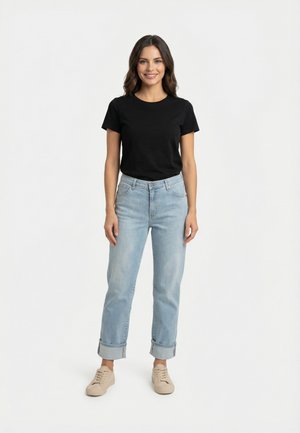 Mujer de pie, vistiendo una camiseta negra de manga corta, vaqueros azul claro remangados y zapatillas beige, sonriendo frente a un fondo blanco liso.