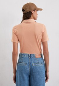 Perzik kleurige polo shirt met korte mouwen, gecombineerd met hoge blauwe spijkerbroek. Pet is van bruin suede. Achteraanzicht benadrukt het merklabel.