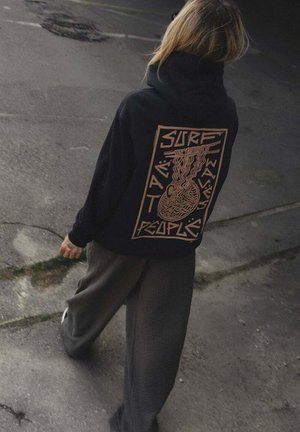 Osoba idąca po chodniku, ubrana w czarną bluzę z kapturem z pomarańczową grafiką i tekstem "Surf Water Waves People" na plecach.