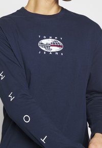 Marinblå långärmad t-shirt med ett broderat globlogga och texten "TOMMY JEANS". Mjuk bomullsmaterial med vita detaljer på ärmarna.