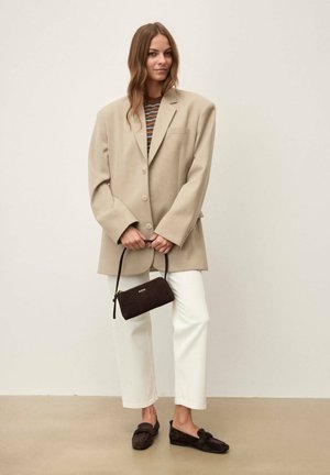 Nő túlméretezett bézs blézert, csíkos felsőt, fehér nadrágot és sötét loafert visel, kezében kis sötét kézitáskát tart, bézs padlón áll, egyszínű háttér előtt.