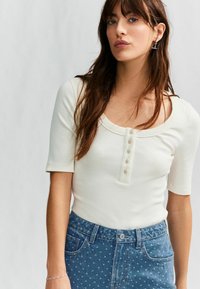 Vit ribbad kortärmad topp med rund halsringning och knappslå, i kombination med blå prickiga jeans.