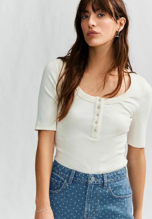 Vit ribbad kortärmad topp med rund halsringning och knappslå, i kombination med blå prickiga jeans.