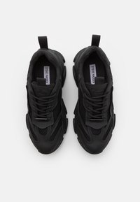 Zapatos deportivos negros con parte superior de malla, diseño texturizado, lengüeta acolchada y suela de goma con contornos prominentes.