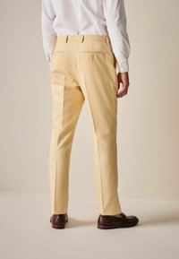 Helle gelbe Hosen aus weichem Stoff, mit einer taillierten Passform, zwei Gesäßtaschen mit Reißverschlüssen und kombiniert mit braunen Schuhen.
