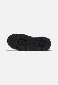 Suela de bota de goma negra con un patrón de relieve texturizado, que presenta surcos en zigzag, un sello de logotipo y un punto verde cerca del talón.