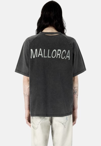 Personne aux longs cheveux foncés vue de dos portant un T-shirt noir délavé avec "MALLORCA" imprimé dans le dos et un jean de couleur claire.
