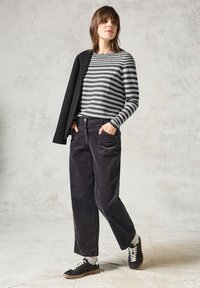 Gestreepte zwart en grijze trui over donkere corduroy broek, met handen in de zakken. Zwarte sneakers met witte veters en een grijze textuur achtergrond.