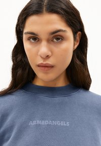 Sudadera azul con cuello acanalado, que presenta un sutil gráfico del logo "ARMEDANGELS" en el centro. Tejido suave y texturizado.