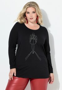 Blondýna v černém dlouhém rukávu s konturou módní postavy z kamínků a v červených kožených kalhotách pózuje před bílou zdí.