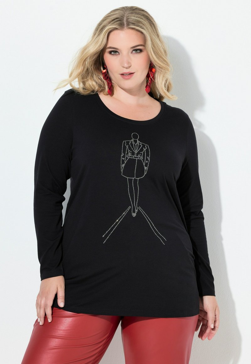 Blondýna v černém dlouhém rukávu s konturou módní postavy z kamínků a v červených kožených kalhotách pózuje před bílou zdí.