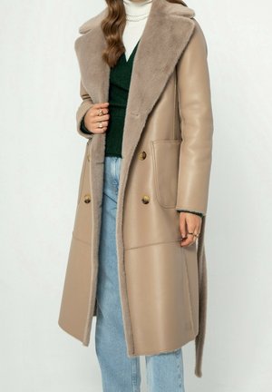 Kvinde iført en lang beige læderjakke med pelskant, grøn sweater, hvid rullekrave, blå jeans og flere guldringe på begge hænder.